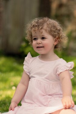 Evoto A young child with curly hair sits on the grass, wearing a light pink dress with ruffled sleeves, looking slightly to the side and smiling—an exquisite moment captured through portrait photography.