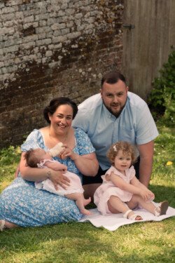 A family of four sits on the grass, captured perfectly in this moment of portrait photography. A woman in a blue dress holds a baby with a bottle, while a man kneels behind a young child in a pink dress. A brick wall and wooden door serve as the backdrop.