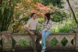 A man and woman sit on a stone railing in a garden, talking and smiling at each other, capturing the charm of the moment through portrait photography.