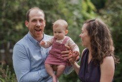 A portrait photography scene captures a man holding a baby while standing next to a woman. They are outdoors with lush greenery in the background. The man is smiling, the baby looks serious, and the woman is laughing.
