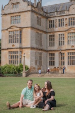 A man, woman, and young girl sit on a lawn in front of a large historic building. The man and woman smile while the girl sits between them, capturing a perfect moment of portrait photography.