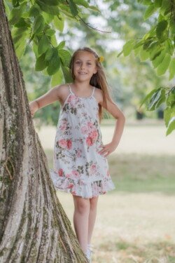 A young girl in a floral dress stands against a tree in a park, her smile captured perfectly for this portrait photography moment.