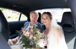 A bride in a wedding dress and veil sits in a car holding a bouquet of flowers next to an older man in a suit. Both are smiling, capturing the joyful spirit often seen at Haselbury Mill Weddings.