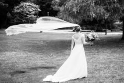 A bride in a white gown stands on grass at Haselbury Mill Weddings, holding a bouquet, with her veil blowing dramatically in the wind.
