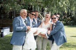 Five men in blue suits and a woman in a white dress smile and laugh outdoors as they hold someone wrapped in white fabric under a tree, capturing the joyful spirit of Haselbury Mill Weddings.