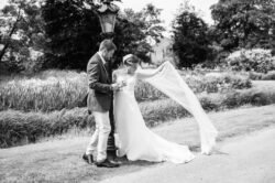 A bride and groom stand outdoors by a lamp post at Haselbury Mill Weddings; the bride holds her long veil up while the groom gently holds her waist in a scenic garden or park setting.