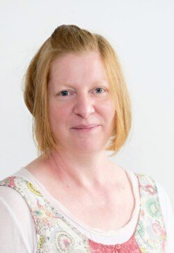 A person with short, light red hair and fair skin, wearing a white shirt with a patterned vest, looks directly at the camera against a plain white background—ideal for corporate photography & video services.