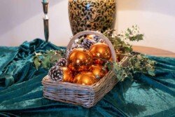 Evoto A wicker basket with shiny gold ornaments, pinecones, and holly sits on a green velvet cloth, reminiscent of the elegant decor seen in Orchardleigh House photos, with a decorative lamp softly glowing in the background.