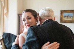 Evoto In a warmly lit room at Orchardleigh House, a bride hugs an older man, both in formal attire. She looks content with a slight smile, the perfect moment captured in these photos.