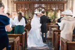 Evoto A couple stands at the altar during a church wedding ceremony, surrounded by guests seated in pews. The scene echoes the elegance of Orchardleigh House photos, with beautiful floral arrangements setting a perfect backdrop.