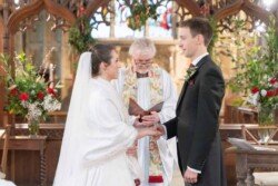 Evoto A couple holding hands during a wedding ceremony at Orchardleigh House, with a priest officiating in a flower-adorned church.
