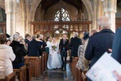 Evoto A bride and groom make their way down the aisle of a church, surrounded by standing guests, capturing a moment as enchanting as any found in Orchardleigh House photos.
