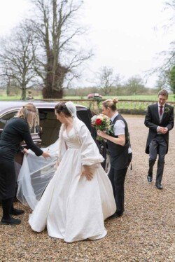 Evoto A bride in a white dress steps out of a car at Orchardleigh House, assisted by two women. A man in formal wear approaches, with one woman holding a bouquet. Trees frame the scene against an overcast sky.