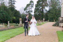 Evoto A bride and groom walk on a gravel path in a garden, surrounded by trees and statues. The bride holds a bouquet, both smiling warmly, echoing memories captured in their Orchardleigh House photos.