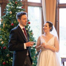 Evoto A couple stands by a decorated Christmas tree. The man, in a suit holding a drink, and the woman, in a white dress with a veil, also holding a drink, are reminiscent of an enchanting Orchardleigh House moment.