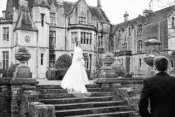 Evoto A bride in a white gown stands on the stone steps of Orchardleigh House, a large historic building, as a person in a suit approaches her, capturing a timeless moment reminiscent of classic wedding photos.
