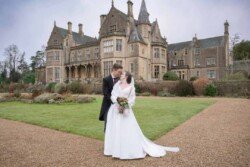 Evoto A bride and groom stand on a gravel path, embracing in front of the majestic Orchardleigh House, its historic facade framed by manicured gardens—a perfect moment captured forever in photos.