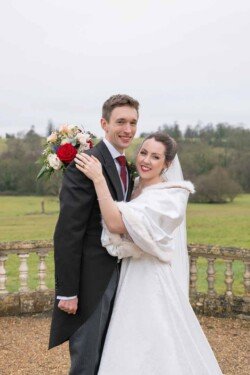 Evoto A couple poses outdoors in formal wedding attire, the woman elegantly holding a bouquet. The rural landscape, reminiscent of Orchardleigh House photos, features trees in the background, adding a timeless charm to the scene.