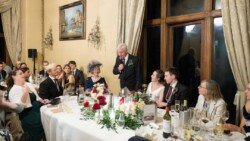 A man in a suit delivers a speech at a formal dinner event, as guests seated around tables adorned with flowers, glasses, and wine bottles create an atmosphere reminiscent of Orchardleigh House photos.