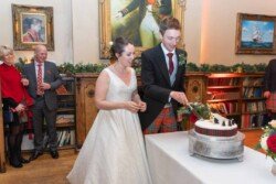 In a book-lined room at Orchardleigh House, a bride and groom cut their wedding cake, surrounded by a few guests. The groom sports tartan pants with his suit, while the bride dazzles in her white dress.