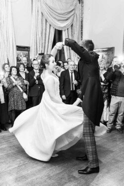 A couple dances in formal attire at an elegant indoor event, surrounded by a crowd of onlookers, capturing the timeless beauty often seen in Orchardleigh House photos.