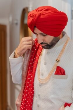 A man wearing a red turban, white traditional attire, and gold necklaces adjusts his red shawl outside the historic Guildhall Bath.