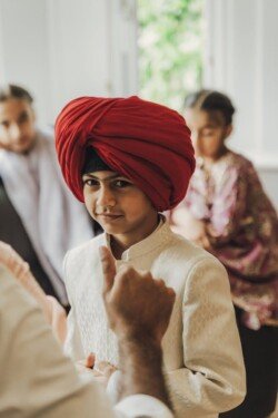 A boy in a white outfit and a red turban looks at the camera while an adult gestures in the foreground. Using Fuji Cameras for weddings, other people are slightly blurred in the background.