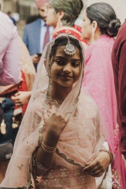 A young girl, dressed in traditional attire with a decorative headpiece and bracelets, smiles amidst a group of people wearing colorful clothing at an outdoor gathering, all captured beautifully by Fuji Cameras for weddings.