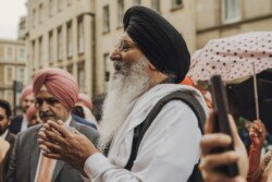 An elderly man in a black turban and white clothing stands among a group of people outdoors, holding his hands together, as if posing for Fuji Cameras for weddings. Beside him, another man in a pink turban stands serenely.