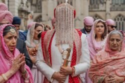 A groom wearing a red and white traditional outfit and a beaded face covering holds a sword. He is surrounded by people in traditional clothing, standing proudly in front of the historic Guildhall Bath.