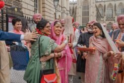 A group of people, dressed in traditional attire, are celebrating in front of the Guildhall Bath. One woman in pink is being hugged as others around her smile and hold festive items.