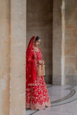 A woman dressed in a red, intricately embroidered traditional outfit stands in the archway of the Guildhall Bath, holding decorative tassels and looking down.
