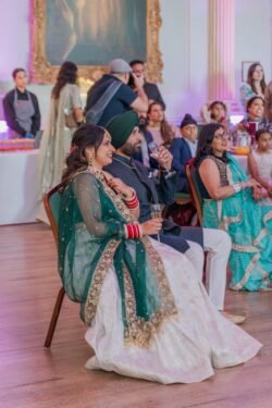 A couple in traditional attire are seated, clapping and smiling, at a formal indoor event at Guildhall Bath. Other guests and servers are visible in the background.