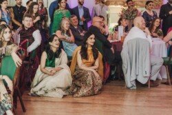 A group of people seated and standing, some smiling and laughing, intently watching something off-camera at an indoor gathering, possibly captured with Fuji Cameras for weddings.