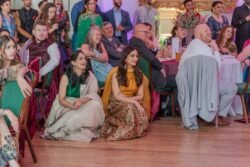 A group of people at an indoor event at Guildhall Bath, many seated and some standing, smiling and attentively watching something. Two women in traditional attire sit on the floor in front of the seated guests.