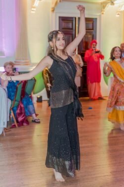 A woman in a black traditional outfit dances barefoot on the wooden floor of the Guildhall Bath, an indoor event space adorned with decor and bustling with people in the background.