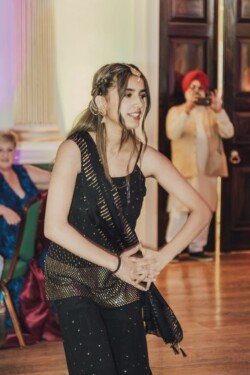 A young woman in a black and gold outfit dances indoors, while an older man in a turban captures the moment in the background using Fuji Cameras for weddings.