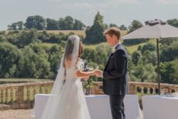 A couple stands outdoors at Orchardleigh House, exchanging rings during their wedding ceremony. The bride wears a white dress and veil, the groom a dark suit. They are surrounded by greenery and a sunny landscape.