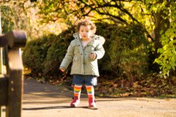A portrait photography moment captures a toddler wearing a green coat, denim skirt, and colorful striped socks standing on a sunlit path, surrounded by lush, green foliage.
