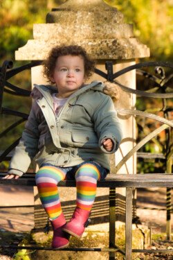 A young child with curly hair sits on an ornate metal bench wearing a green coat, rainbow-striped tights, and red boots, looking slightly to the left. This charming instance of portrait photography is set against a sunlit outdoor background.