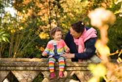 In a charming example of portrait photography, a woman and a young child sit on a stone wall in a park. The child, dressed in a colorful outfit, beams with joy as the woman leans in to chat with them.
