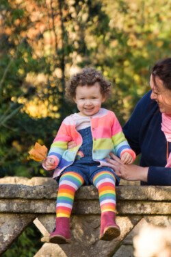 Captured through the lens of skilled portrait photography, a child with curly hair and rainbow tights sits on a stone ledge holding a leaf, while an adult in a blue jacket and pink scarf stands beside them.