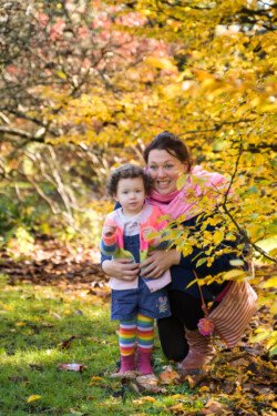 An adult and a child stand in a park with colorful autumn leaves, perfect for portrait photography. The adult is kneeling and hugging the child from behind. The child is wearing a denim dress and rainbow-striped leggings.