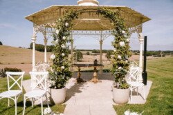 An outdoor gazebo at Hinton Park decorated with white flowers and greenery, featuring white chairs and a small table, set against a backdrop of an expansive field under a clear sky, perfect for Croftview weddings.