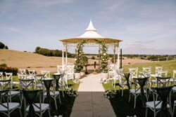 An outdoor wedding setup at Croftview Weddings in Hinton Park features a white gazebo adorned with greenery, with rows of white chairs on either side of a stone aisle, set against a backdrop of open fields and a clear sky.