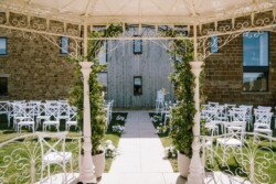 Outdoor wedding venue at Croftview Weddings in Hinton Park features white chairs arranged in rows, a central aisle adorned with greenery and flowers under a wrought-iron gazebo, with a rustic building exterior visible in the background.