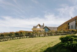 An open grassy field at Hinton Park slopes gently upward toward a house with multiple rooflines and a mix of modern and rustic architecture, bordered by a wooden fence and shrubs. The sky is clear with wispy clouds, making it an idyllic setting for Croftview weddings.
