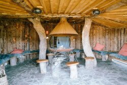 A rustic wooden gazebo with a central fire pit, tree trunk support columns, stone flooring, bench seating with cushions, and a ceiling with exposed wooden beams offers the perfect setting for Croftview weddings at Hinton Park.