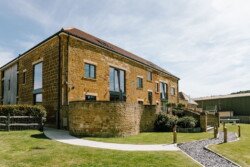 A large stone building with modern windows and a red-tiled roof, Hinton Park offers the perfect setting for Croftview weddings. Surrounded by a well-maintained lawn and a paved pathway on a sunny day, it's an idyllic venue.