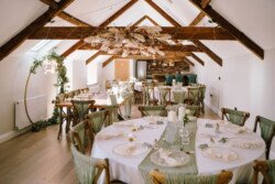 A rustic wedding reception setup at Hinton Park features wooden beams, round tables with white linens, green table runners, and elegant table settings. A floral arrangement hangs from the ceiling, creating the perfect backdrop for Croftview weddings.